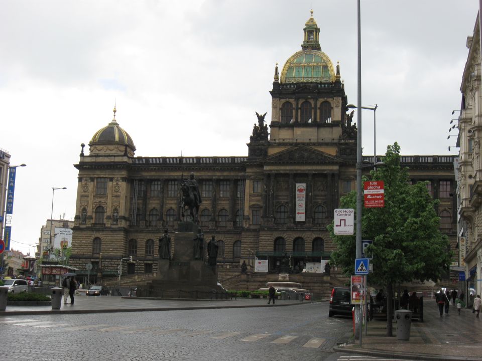 Historic building with a dome and a statue in a city square, with a few cars and pedestrians visible