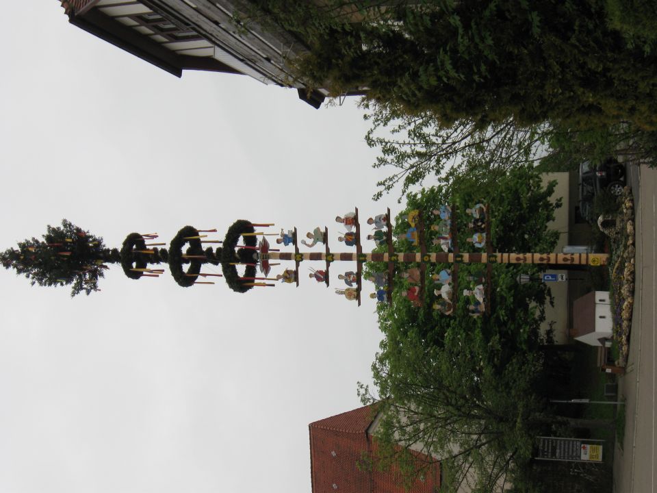 Maypole with figures and wreaths, topped with a tree, against a cloudy sky