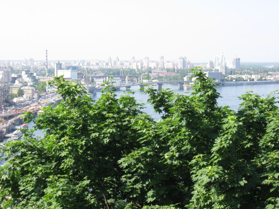 City skyline across a wide river, with a bridge in the middle distance and trees in the foreground