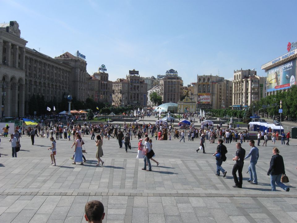 Crowded city square with large buildings and many people walking under a clear blue sky. A watermark reads SUPERMOTORS