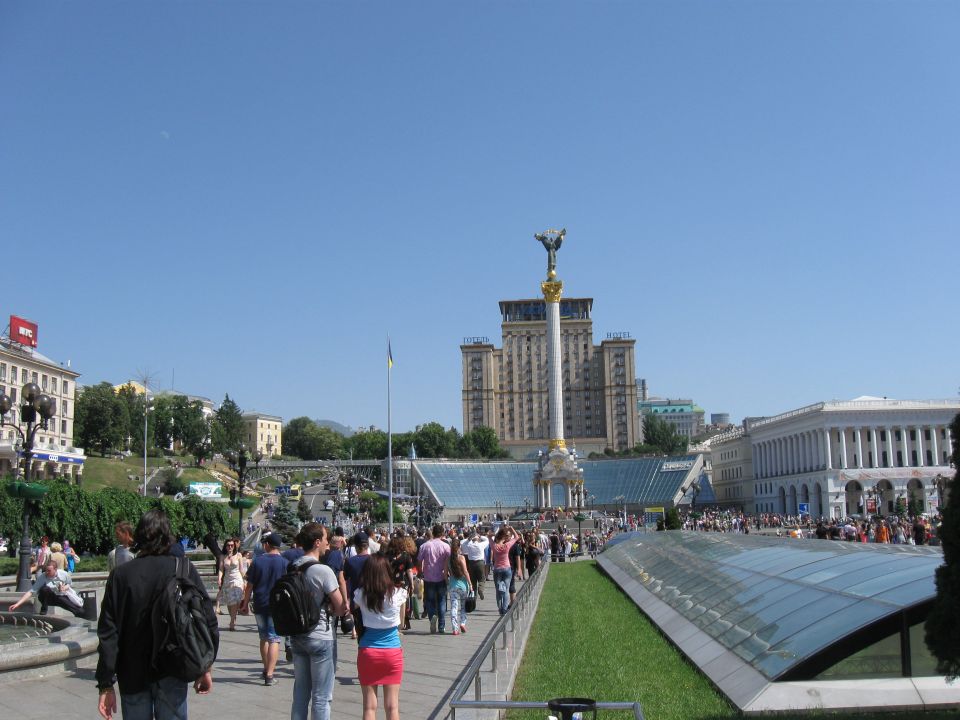 City square with a tall monument topped by a statue, surrounded by buildings and a crowd of people walking