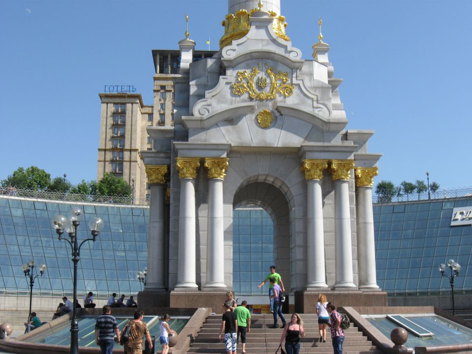 Monumental archway with golden accents and columns, people walking on stairs below. Hotel building in background
