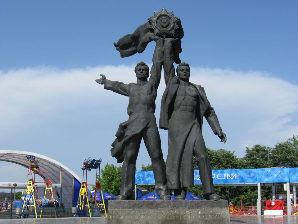 Monumental sculpture of two men holding a star against a blue sky with clouds. A banner is visible behind them