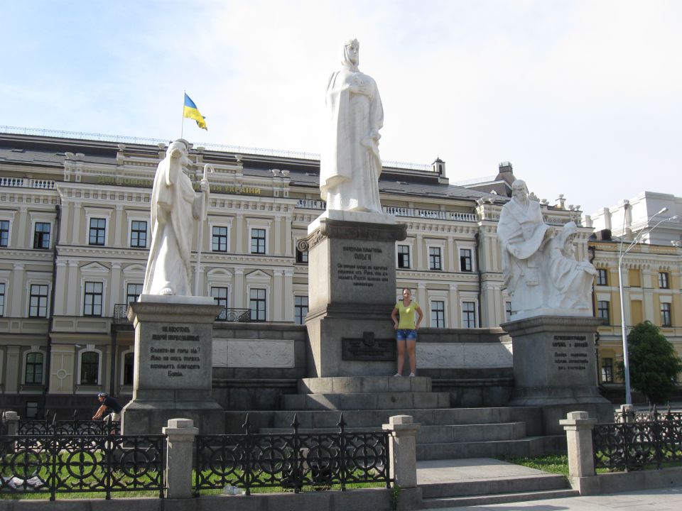 Monument with white statues and a Ukrainian flag, in front of a light-colored building. A person stands on the monument's steps