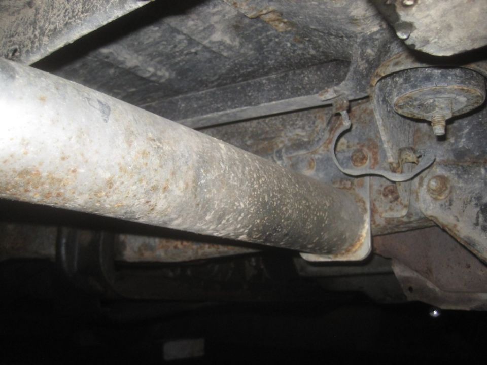 Underside view of a vehicle's exhaust pipe and frame. Shows rust and some mounting hardware