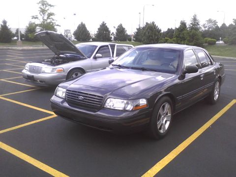 2003 Ford Crown Victoria - A dark-colored Ford Crown Victoria is parked in front of a silver one with its hood up.