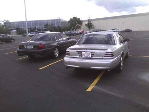 2003 Ford Crown Victoria - Two Ford Crown Victorias are parked side-by-side in a parking lot with a large building in the background.