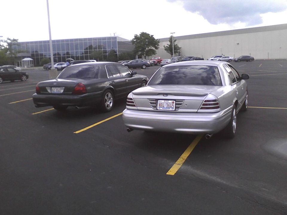 Two Ford Crown Victorias parked in a lot. One is silver, the other is dark green. The silver one has a license plate that reads NBL 3645