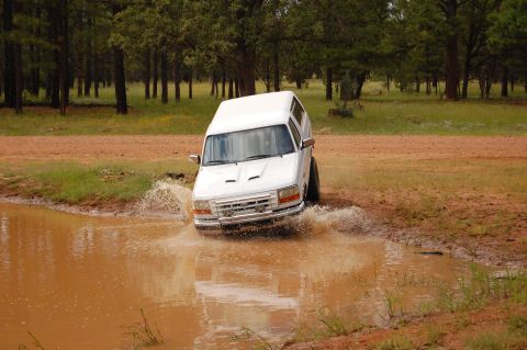 1996 Ford Bronco - A white SUV navigates a muddy puddle in a wooded area.