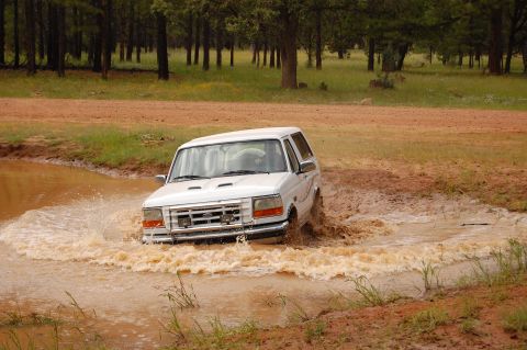 1996 Ford Bronco - A white 1996 Ford Bronco navigates through a water-filled ditch on a dirt road.