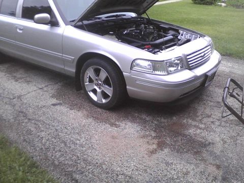 2003 Ford Crown Victoria - The front of a silver 2003 Ford Crown Victoria is shown with the hood open, revealing the engine compartment.