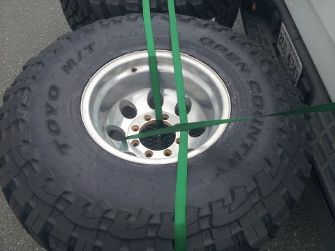 1988 Ford Bronco - Spare Toyo Open Country M/T tires are secured with green straps to the back of a white vehicle, with a partial license plate visible.