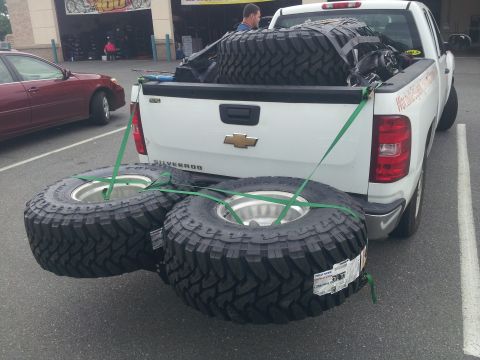 1988 Ford Bronco - A white Chevrolet Silverado pickup truck is loaded with several large off-road tires, with two tires strapped to the rear bumper.
