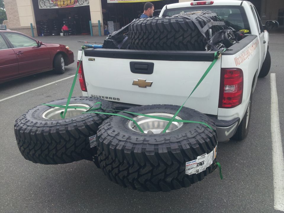White pickup truck bed loaded with large off-road tires, two secured to the rear bumper