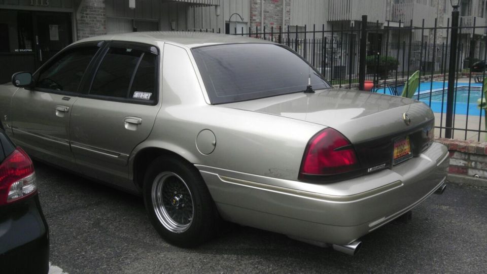 Silver 2000 Mercury Grand Marquis sedan, rear 3/4 view, tinted windows, aftermarket wheels, parked next to a dark car