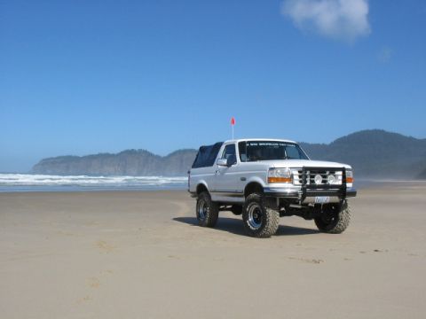 1995 Ford Bronco - A lifted 1995 Ford Bronco sits on a sandy beach with the ocean and coastal hills in the background under a clear blue sky.