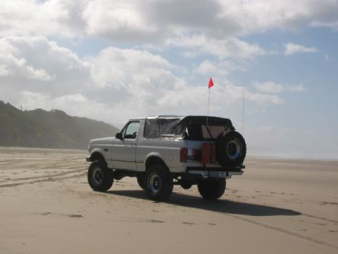 1995 Ford Bronco - A white Ford Bronco with a soft top and spare tire is parked on a sandy beach under a cloudy sky.