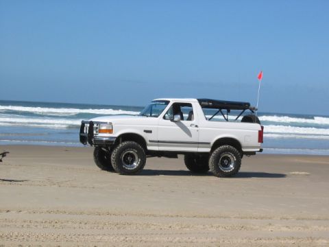 1995 Ford Bronco - A white 1995 Ford Bronco is parked on a sandy beach with the ocean in the background.