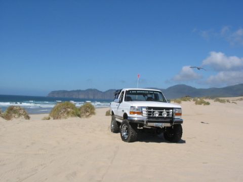 1995 Ford Bronco - A lifted pickup truck is parked on a sandy dune near the ocean on a clear day.