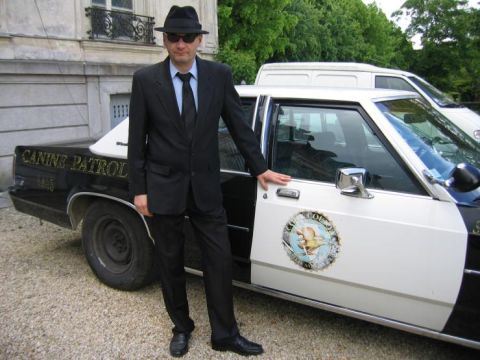 1991 Ford LTD Crown Victoria - A man dressed as a police officer stands next to a marked patrol car, with a white van visible in the background.