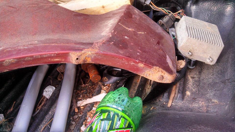 rusted red car fender section with wires and a gray box attached, next to a green soda bottle