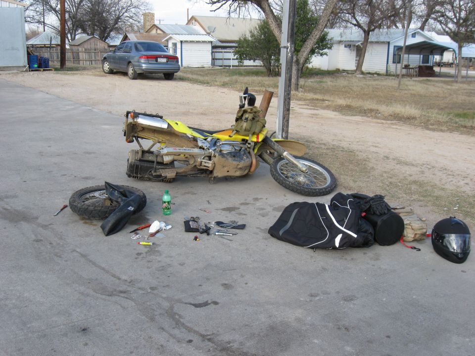 Dirt bike on its side with tools, a helmet, and bags laid out on the pavement