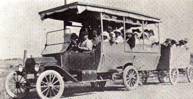 Bus with canvas roof and open sides, carrying passengers. Visible front grille, headlights, and spoked wheels