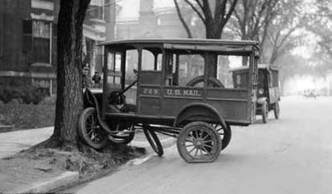 1909 Ford Model T - A U.S. Mail truck with a flat tire is parked by a tree, with another truck visible in the background on a tree-lined street.