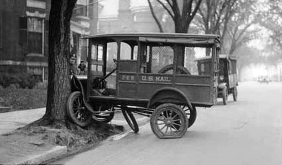 Side view of a U.S. Mail truck with flat tire parked next to a tree. Another truck is visible behind it