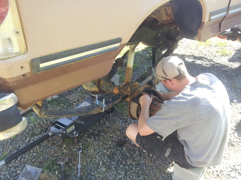 Guy working on the rear axle and suspension of a tan truck. It looks like a Ford Bronco