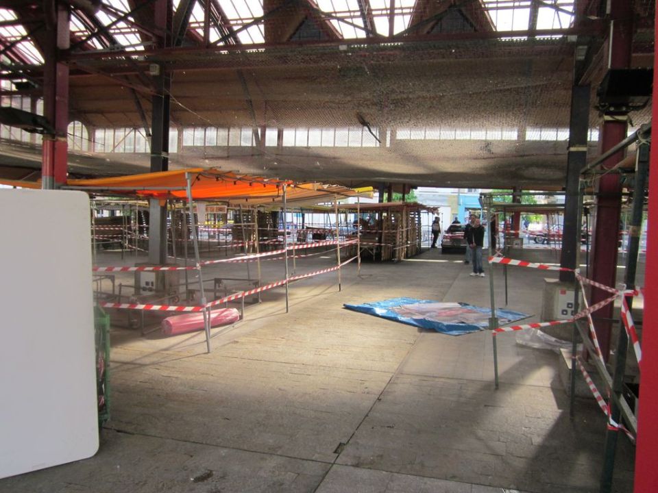 Market stalls under a covered structure with a red car visible in the distance