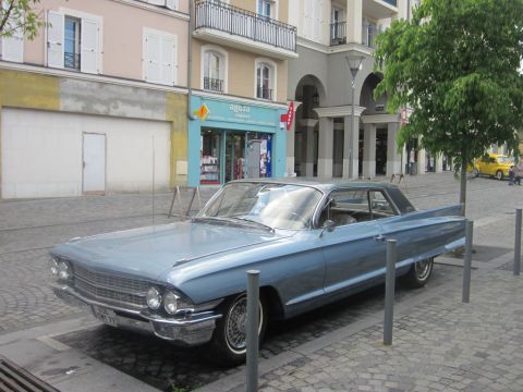 1991 Ford LTD Crown Victoria - A light blue 1960 Cadillac sedan is parked on a cobblestone street in front of shops.