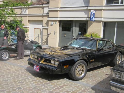 1991 Ford LTD Crown Victoria - A black 1977 Pontiac Firebird Trans Am is parked on a cobblestone street, with other vehicles and people nearby.