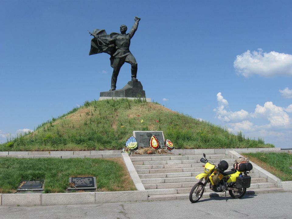 Yellow dual-sport motorcycle parked on pavement in front of a monument with a statue