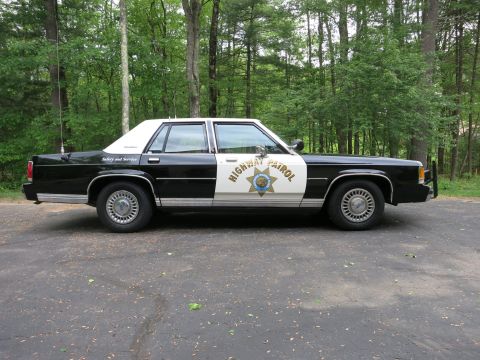 1991 Ford LTD Crown Victoria - black-and-white police cruiser marked Highway Patrol with a gold star door shield, shown in full side profile in a parking lot.