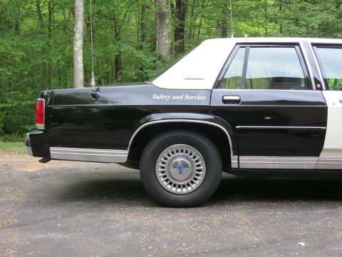 1991 Ford LTD Crown Victoria - Close rear-quarter view of a black-and-white police sedan with a Ford-logo hubcap and gold Safety and Service lettering.