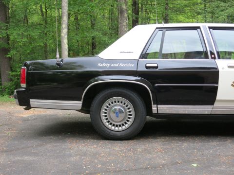 1991 Ford LTD Crown Victoria - Rear-quarter panel of a black-and-white police cruiser bearing white Safety and Service lettering and a Ford-emblem wheel cover.
