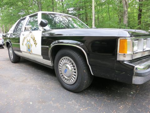 1991 Ford LTD Crown Victoria - Low front-quarter angle of a black-and-white Highway Patrol sedan showing amber turn signal, Ford wheel cover, and door star.