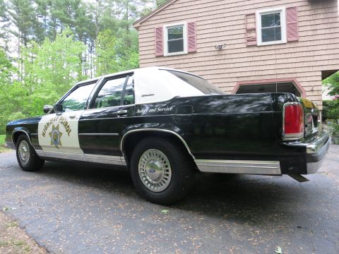 1991 Ford LTD Crown Victoria - Rear-quarter view of a black-and-white Highway Patrol cruiser parked beside a shingled house with Safety and Service lettering.