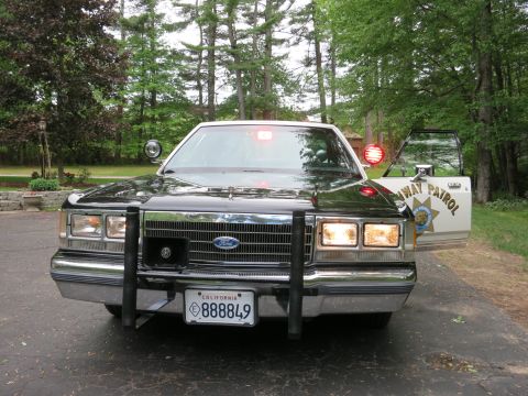 1991 Ford LTD Crown Victoria - Front of a black-and-white Ford sedan with push bumper and lit headlights, the open door marked 'Highway Patrol'.