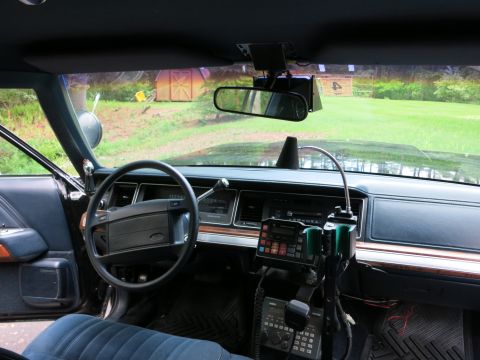 1991 Ford LTD Crown Victoria - Ford car dashboard and steering wheel with rearview mirror, whip antenna and mounted radio gear; a barn is visible outside.