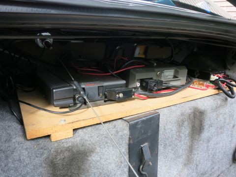 1991 Ford LTD Crown Victoria - Vehicle trunk shelf holding black radio amplifier units and antenna cabling mounted on a wood platform above carpet.