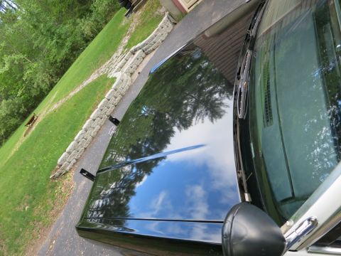 1991 Ford LTD Crown Victoria - Glossy black car trunk and rear glass reflecting sky and trees, with a stacked-stone retaining wall in the background.