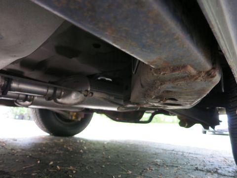 1991 Ford LTD Crown Victoria - Undercarriage view showing a rust-stained floor pan, exhaust resonator pipe and a rear tire over pavement.
