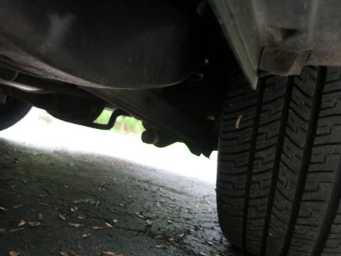 1991 Ford LTD Crown Victoria - Underside view beside a rear tire showing the exhaust pipe, chassis members and wet pavement.