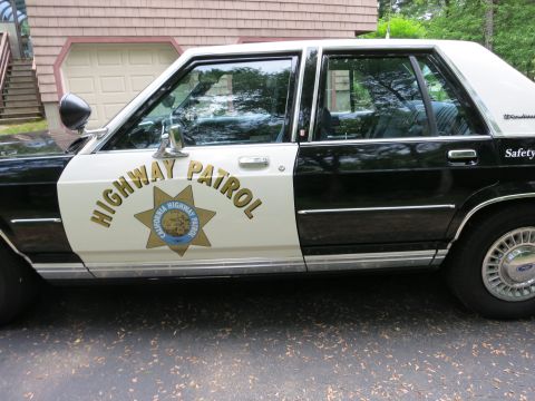 1991 Ford LTD Crown Victoria - Driver-side view of a black-and-white California Highway Patrol Ford sedan with gold star emblem and chrome wheel covers, parked by a house.
