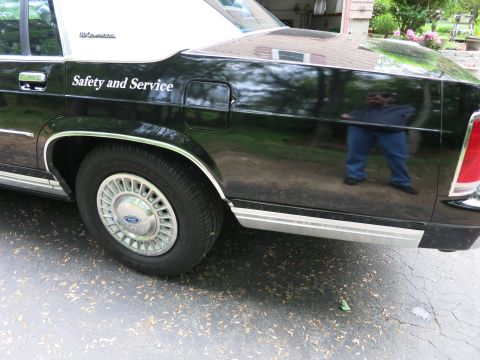1991 Ford LTD Crown Victoria - Rear quarter of a black police sedan reading 'Safety and Service' with a Ford-badged chrome wheel cover, photographer reflected in the pain…