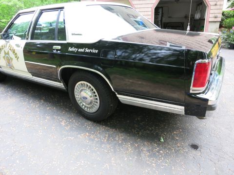 1991 Ford LTD Crown Victoria - Rear three-quarter view of a black-and-white CHP police sedan showing taillights, 'Safety and Service' lettering and a Ford wheel cover.