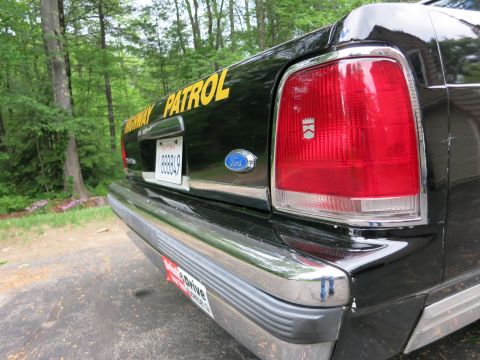 1991 Ford LTD Crown Victoria - Low-angle rear corner of a black police car showing yellow 'Highway Patrol' lettering, a Ford-badged trunk and red taillight.