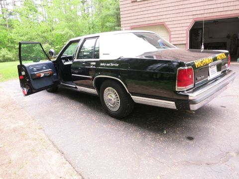 1991 Ford LTD Crown Victoria - Rear three-quarter view of a black-and-white CHP police sedan with the rear door open revealing the blue back seat, by a garage.